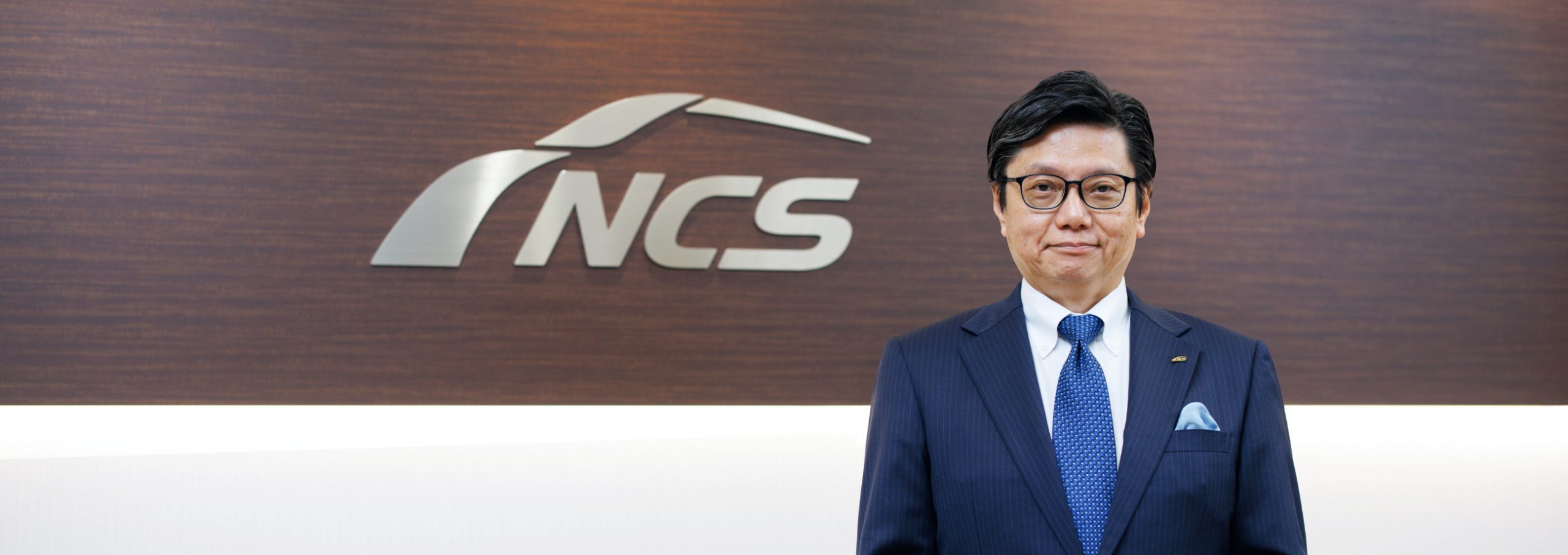 NCSの看板の前に立つ代表取締役社長の写真