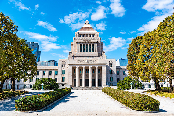 青空の下、並木道の先に正面入口を構える国会議事堂の建物全景